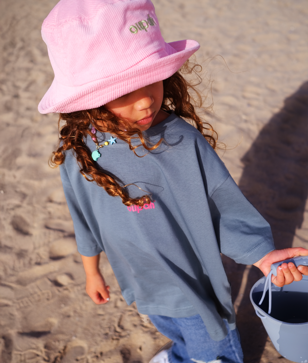 Little girl in a pink bucket hat wearing the Oupah Clockwork Tee on the beach