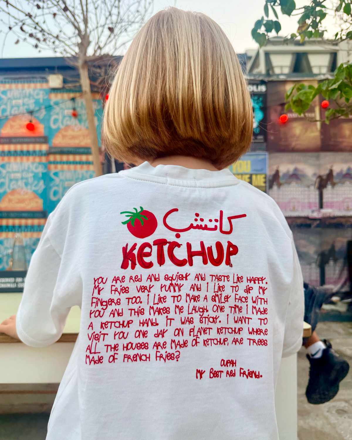 Back of the Oupah Ketchup Tee with red Arabic and English Ketchup print, worn by a child