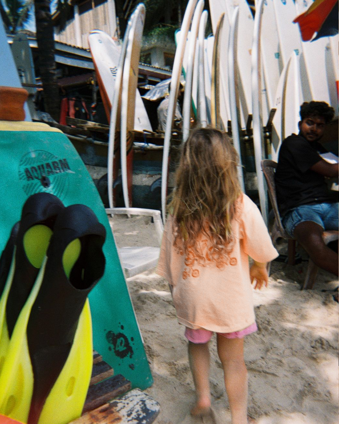 Little girl on the beach wearing the blush Oupah Wheels Feels Tee, showing the orange print on the back